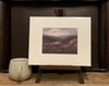 A photo of a Giclée print from Judith Levin original oil painting After Rain, depicting the Yorkshire Moors, on a mini easel next to a mug.