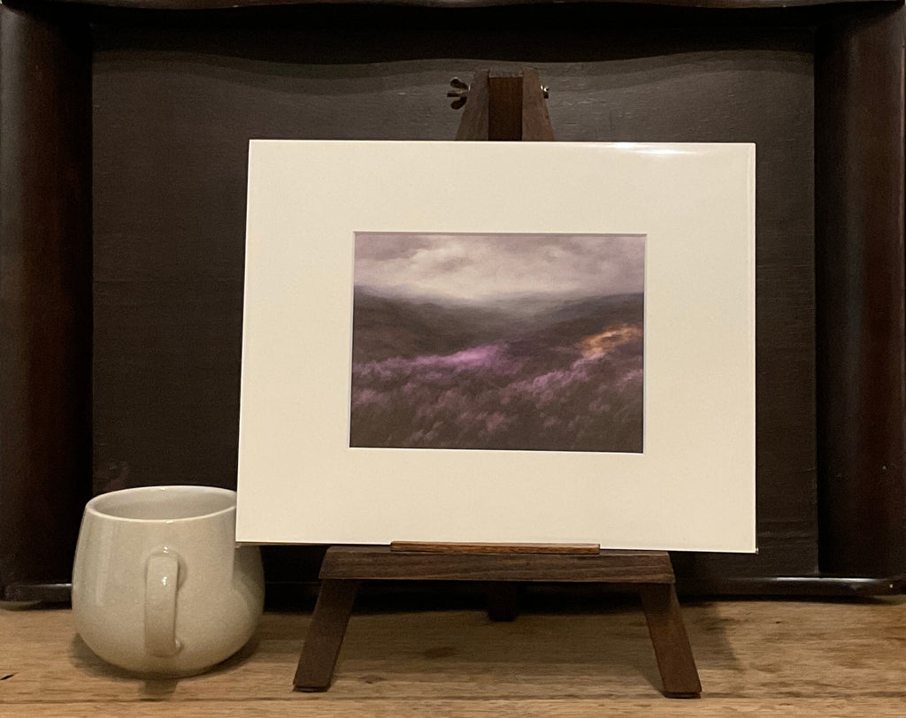 A photo of a Giclée print from Judith Levin original oil painting After Rain, depicting the Yorkshire Moors, on a mini easel next to a mug.