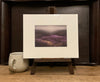 A photo of a Giclée print from Judith Levin original oil painting Heather Valley, depicting heather moorland, on a mini easel next to a mug.